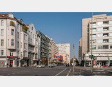 Straßenszene in einer Stadt mit mehrstöckigen Wohn- und Geschäftshäusern auf beiden Seiten, teils mit Jugendstil-Fassaden. Im Hintergrund größere Bürogebäude, darunter eines mit dem Logo "Allianz", und Verkehrsampeln sowie Autos auf der Straße.