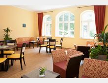 Ein gemütlicher Aufenthaltsraum mit gelb gestrichenen Wänden, großen Fenstern mit Rundbögen und roten Vorhängen. Der Raum ist mit Tischen, Stühlen mit gelben Polstern und rotem gemustertem Stoff sowie Pflanzen dekoriert.