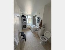 Schmales Badezimmer mit weißen Fliesen, Waschbecken mit Spiegel, Toilette und Badewanne unter einem großen Fenster. Am Fenster steht eine gestapelte Waschmaschine mit Trockner; links ist ein Heizkörper und ein kleiner Hocker, rechts ein dunkler Fliesenstreifen als Akzentwand.