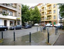 Straßenszene in einer Wohngegend mit mehrstöckigen Gebäuden im Altbaustil, darunter Geschäfte im Erdgeschoss. Pflastersteine, geparkte Autos, Motorräder sowie Bäume und Straßenschilder prägen den Bereich.