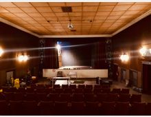 Innenraum eines kleinen Theatersaals mit Reihen gepolsterter Stühle, einer Bühne im Hintergrund und dekorativen Holzelementen an Decke und Wänden. Rechts und links der Bühne befinden sich Vorhänge und der Schriftzug „Alhambra“ ist sichtbar.