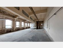 Verlassener, leerer Raum in einem Industriebau mit abblätternder Farbe an Wänden und Decke, mehreren großen Fenstern entlang einer Wand, sichtbaren Kabelrohren und abgewittertem Betonboden.