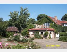 Backsteinhaus mit Ziegeldach, teilweise von Efeu bewachsen, umgeben von einem Garten mit blühenden Rosen und Bäumen; im Vordergrund ein gepflasterter Hof, im Hintergrund dichte grüne Vegetation.