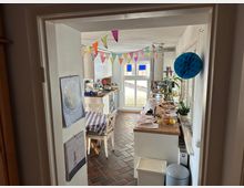 Blick in eine helle Küche mit dunklem Fliesenboden und einer langen Arbeitsfläche aus Holz. Ein Tisch mit blau-weiß-karierter Tischdecke steht in einer Ecke, an der Wand hängen bunte Wimpel und Deko-Elemente.