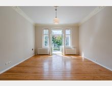 Leerer, heller Altbau-Wohnraum mit breiten Holzdielen, hohen Decken mit Stuck und zentraler Hängelampe. Am Ende des Raums befinden sich hohe Fenster und eine geöffnete Balkontür, flankiert von Heizkörpern, mit Blick ins Grüne.