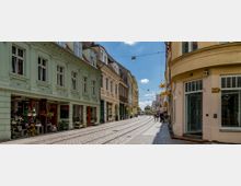 Eine gepflasterte Straße in einer Altstadt mit Straßenbahnschienen, flankiert von historischen Gebäuden mit pastellfarbenen Fassaden. Auf der linken Seite befinden sich Geschäfte mit Blumen und Dekorationen, während Passanten entlang der Straße spazieren.