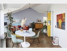 Offener Wohnraum mit hellblauen Wänden, einem Esstisch aus weißem Marmor, umgeben von pastellfarbenen Stühlen. Im Hintergrund befinden sich ein rotes Sofa, Pflanzen, ein Holzschrank und eine gelbe Bücherwand, ergänzt durch Dekoration wie eine Discokugel und Wandkunst.