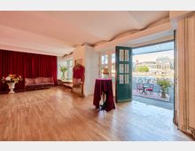 Großer, heller Innenraum mit Holzparkett, hohen weißen Decken und burgunderroten Vorhängen, ausgestattet mit einem roten Sofa, einem hohen Spiegel und dekorativen Blumenarrangements. Eine offene türkisfarbene Glastür führt zu einer Terrasse am Wasser mit Café-Tischen; im Hintergrund sind moderne Gebäude zu sehen.