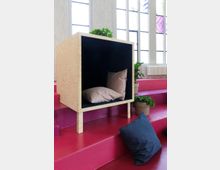 Ein minimalistisches Interieur mit einer Holzkonstruktion, die ein würfelförmiges Sitzmöbel mit schwarzen Innenwänden und Kissen darstellt, auf pinken Stufen platziert. Im Hintergrund sind große Fenster mit Gitterrahmen, Pflanzen in Töpfen und ein Heizkörper zu sehen.