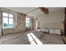 Ein leerer, renovierungsbedürftiger Raum mit abblätternden Tapeten, freigelegtem Mauerwerk und einem beschädigten Holzfußboden. Die großen Fenster lassen viel Tageslicht herein, und mehrere alte Türen lehnen an einer Wand.