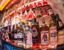 Nahaufnahme eines beleuchteten Barregals mit einer Vielzahl von alkoholischen Getränkeflaschen, darunter Spirituosen wie Rum, Whiskey und Likör. Die Flaschen sind in mehreren Reihen angeordnet, und im Hintergrund sind weitere Flaschen auf Glasregalen sichtbar.