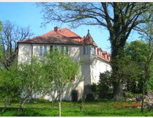 Historisches Gebäude mit hellen Fassaden und roten Ziegeldächern, umgeben von Bäumen und Grünflächen. Im Vordergrund stehen Bäume und Büsche, während der blaue Himmel im Hintergrund sichtbar ist.