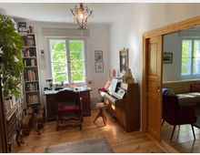 Ein heller Arbeitsraum mit Holzboden, einem Schreibtisch vor einem großen Fenster mit Blick ins Grüne, einer Bücherwand links und einem Klavier rechts. Im angrenzenden Raum, erkennbar durch eine offene Holztür, befindet sich ein gelbes Sofa und ein Sessel.