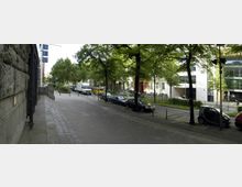 Straßenszene mit breitem Gehweg und parkenden Autos entlang der Straße, gesäumt von Bäumen und Gebäuden im Hintergrund, darunter moderne Glasfassaden und ältere, helle Fassaden. Links ist eine grob strukturierte Steinmauer zu sehen, während rechts Fahrräder und Fußgänger den Gehweg nutzen.
