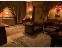 Gemütlicher Raum mit gedämpfter Beleuchtung, eingerichtet mit dunkelbraunen Ledersofas, Holzmöbeln und Teppichen mit orientalischen Mustern. Die Wände sind mit gelben Vorhängen und Dekorationen wie Bildern und kleinen Lampen gestaltet.