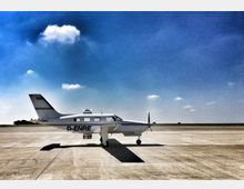 Ein kleines Propellerflugzeug steht auf einer asphaltierten Landebahn unter klarem, blauem Himmel mit einigen kleinen Wolken. Im Hintergrund ist eine flache Landschaft ohne sichtbare Gebäude.