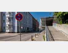 Straßenansicht mit mehrstöckigen Wohnhäusern auf der linken Seite und einer modernen Fußgängerüberführung mit Treppe auf der rechten Seite. Im Vordergrund befinden sich ein Halteverbotsschild und eine Tempo-20-Zone-Beschilderung.