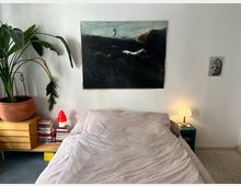 Minimalist bedroom with a low bed and light gray bedding against a white wall, featuring a large dark landscape painting above the headboard. A large potted plant sits on a wooden cabinet beside stacks of books and a small red lamp, with a bedside table holding a lit lamp on the other side.