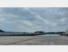 Industriehafen mit einer betonierten Uferfläche, Lagerhallen, Materialhaufen und einem Anlegesteg entlang des Wassers; im Hintergrund sind Bäume und ein bewölkter Himmel sichtbar.