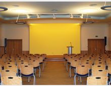Ein Veranstaltungsraum mit Reihen aus Holzstühlen, einer kleinen Bühne mit einem Rednerpult und einem Mikrofon. Die Rückwand der Bühne ist in Gelb gehalten, und der Raum verfügt über eine abgehängte Decke mit kreisförmigen Lampen sowie Holztüren an den Seiten.