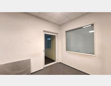 Kleines Büro oder Raum mit weiß gestrichenen Wänden, grauem Bodenbelag und Rasterdecke. Rechts ein Fenster mit Jalousien, links eine Glastür, die in einen angrenzenden Raum führt.