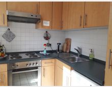 Moderne Küche mit Holzschränken, weiß gefliester Rückwand und Arbeitsfläche aus dunkelgrünem Material. Sichtbar sind ein Edelstahlherd mit vier Kochplatten, eine Dunstabzugshaube, eine Spüle mit Wasserhahn und verschiedene Küchenutensilien wie Schneidebretter und ein Wasserkocher.