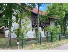 Zweistöckiges Wohnhaus mit weißer Fassade, dunkelbraunen Holzbalkonen und einem roten Ziegeldach, umgeben von Bäumen und einem grünen Metallzaun. Im Vordergrund befindet sich ein gepflasterter Gehweg.