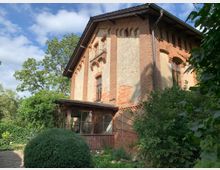 Zweistöckiges Backsteinhaus mit teilweise abgebröckeltem Putz, umgeben von dichtem Garten mit Sträuchern und Bäumen. Im Vordergrund befindet sich ein geschnittener Busch und ein gepflasterter Weg, der zum Haus führt.