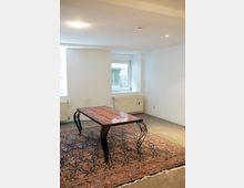 Ein einfacher Innenraum mit weißen Wänden, einem Fenster mit Tageslicht und Heizkörpern. Im Mittelpunkt steht ein verzierter Tisch mit Metallbeinen auf einem großen, gemusterten Teppich, der auf einem Betonboden liegt.