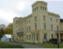 Gelb gestrichenes historisches Gebäude im neoklassizistischen Stil mit rechteckigen Türmen, großen Fenstern und einem dekorativen Balkon; umgeben von einer Rasenfläche und Herbstbäumen. Daneben steht ein großes weißes Zelt.