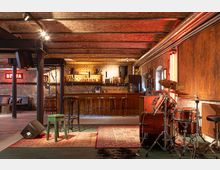 Eine rustikale Bar mit Backsteinwänden und Holzbalkendecke, ausgestattet mit einer langen Theke, Barhockern und Regalen voller Getränkeflaschen. Im Vordergrund stehen ein Schlagzeug, ein Holzhocker und ein Mikrofon auf einem Teppich, was auf eine Bühne für Live-Musik hindeutet.