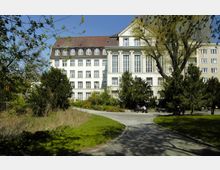 Historisches Gebäude mit heller Fassade, großen Fenstern und Säulen, umgeben von einem kleinen Park mit Gehwegen, Bäumen und gepflegten Grünflächen. Im Vordergrund sind Sitzbänke und eine Gruppe von Menschen erkennbar.