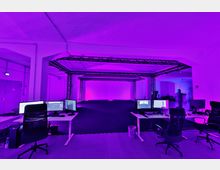 Arbeitsraum mit mehreren Schreibtischen und Computermonitoren im Vordergrund, umgeben von einer großen offenen Fläche mit schwarzen Bodenmatten und einem metallenen Gerüstsystem unter lila Beleuchtung. Die Umgebung wirkt technisch und für Studio- oder Produktionsarbeiten eingerichtet.