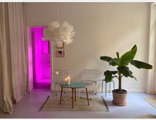 Minimalistisch eingerichtetes Wohnzimmer mit heller Wand, einer Bananenpflanze in einem Topf, zwei weißen Klappstühlen, einem kleinen runden Tisch mit brennenden Kerzen und einem hellen Teppich. Im Hintergrund führt eine Tür zu einem Raum mit pink-violetter Beleuchtung.