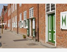 Straßenszene mit einer Reihe historischer Backsteinhäuser, deren Fensterläden und Türen in Grün und Weiß gestrichen sind. Der Gehweg besteht aus Pflastersteinen, und im Hintergrund sind ein Sonnenschirm und Laternen sichtbar.