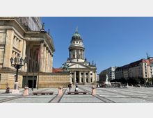 Platz mit Blick auf historische Gebäude, darunter ein Gebäude mit Säulen und Kuppel (Französischer Dom), flankiert von weiteren klassischen und modernen Gebäuden; im Vordergrund gepflasterter Platz mit Bänken und Laternen, im Hintergrund eine Statue.
