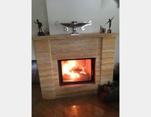 Ein Kamin mit beige-gemaserter Steinverkleidung, in dem ein Feuer brennt. Auf dem Kaminsims stehen eine silberne Schale und zwei kleine Skulpturen, daneben liegt ein Korb mit Holzscheiten auf dem Boden.