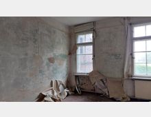 Ein verlassener Raum mit abblätternder Tapete und freiliegenden, beschädigten Wänden. Zwei große Fenster mit Holzrahmen lassen Tageslicht herein, während der Boden und die Heizkörper sichtbar verfallen wirken.