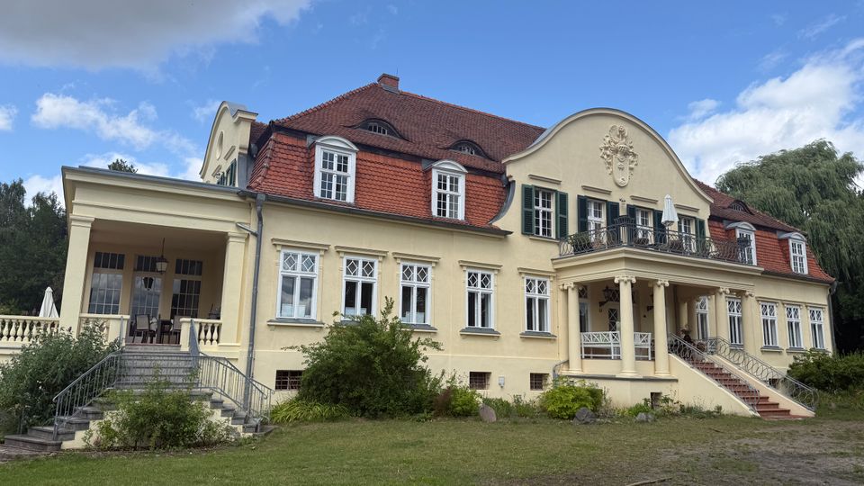 Großes, hellgelbes Herrenhaus mit rotem Ziegeldach und Gauben, symmetrischer Fensterfront sowie einem Balkon über einem Säulenportikus. Links befindet sich eine überdachte Terrasse mit breiter Treppe; davor liegt eine Rasenfläche mit Sträuchern und Bäumen.
