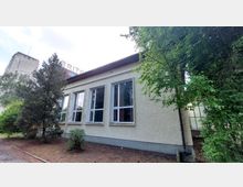 Ein einstöckiges Gebäude mit heller Putzfassade und großen, rechteckigen Fenstern, umgeben von Bäumen und Sträuchern. Im Hintergrund ist ein größeres, teilweise sichtbares Gebäude mit einem Schriftzug zu erkennen.