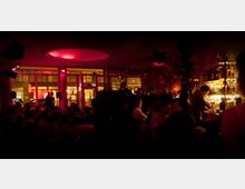 Ein dunkler Innenraum einer Bar mit roter Beleuchtung. Im Vordergrund sitzen und stehen Menschen, während an der rechten Seite eine beleuchtete Theke mit Flaschen sichtbar ist. Im Hintergrund sind große Glasfenster zu sehen, die den Blick auf eine beleuchtete Straße ermöglichen.