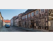 Schmale Kopfsteinpflasterstraße mit historischen Fachwerkhäusern, darunter eines mit sichtbaren Holz- und Putzschäden. Am Straßenrand parken mehrere Autos, und im Hintergrund sind modernere Gebäude mit roten Ziegeldächern zu sehen.