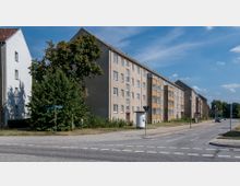 Mehrgeschossiges Wohngebäude aus schlichten, verputzten Fassaden mit schrägen Dächern und mehreren Fenstern, entlang einer ruhigen Straße mit Gehwegen und Straßenlaternen. Ein Baum und einige Büsche befinden sich vor dem Gebäude, im Hintergrund sind weitere Wohnhäuser und Grünflächen sichtbar.