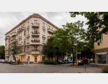 Ecke eines Wohnhauses im Altbaustil mit vier Etagen, hellbeige Fassade und Balkonen, umgeben von Bäumen und parkenden Autos auf einer gepflasterten Straße. Im Erdgeschoss sind Geschäfte erkennbar, darunter ein Laden mit gelber und rosafarbener Gestaltung.