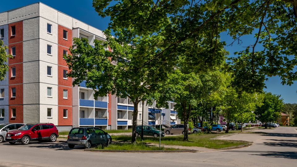 Mehrstöckiges Wohnhaus mit rot-weiß-grauer Fassade und Balkonen mit blauen Brüstungen, an einer ruhigen Straße. Vor dem Gebäude stehen geparkte Autos, und hohe Bäume säumen Gehweg und Grünstreifen.