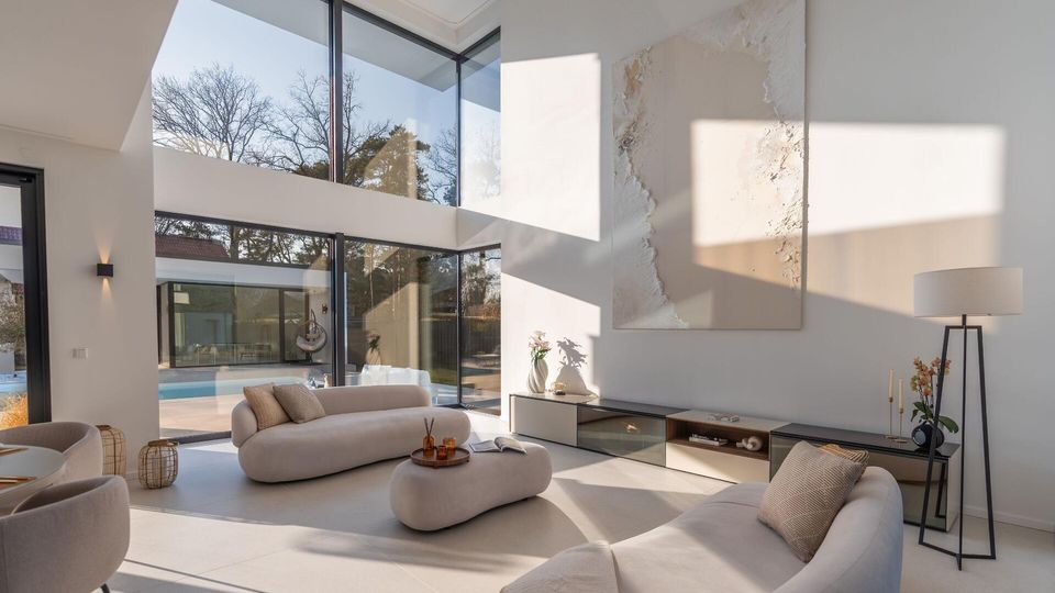 Modernes, doppelgeschossiges Wohnzimmer mit bodentiefen Glasfronten und großer Fensterfront, die auf eine Terrasse mit Pool und Garten blickt. Helle, minimalistische Einrichtung mit niedrigen Sofas, großformatigem Wandbild, flachem Sideboard und Stehlampe in neutralen Farbtönen.