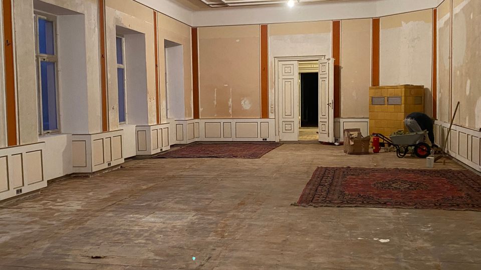Großer, weitgehend leerer Saal in einem Altbau mit hohen Fenstern, Wandvertäfelung und dekorierter Kassettendecke; der Holzboden ist abgenutzt und zwei Teppiche liegen aus. Im Hintergrund steht eine offene Doppeltür, rechts sind Renovierungsmaterialien und ein Kachelofen zu sehen.