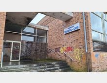 Eingangsbereich eines Backsteingebäudes mit Betonvordach und breiten Stufen, teils mit Graffiti und verwitterten Oberflächen. An der Fassade hängt ein Schild „Restaurant THEATER • CAFÉ“, daneben große Fensterflächen und teils vernagelte Öffnungen.