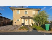 Zweistöckiges, beige verputztes Einfamilienhaus mit dunklem Satteldach und kleiner überdachter Eingangsnische mit weißer Haustür. Vorn liegen eine gepflasterte Einfahrt, ein schmaler Gartenweg mit Beeten und ein kleiner Rasen mit Baum; rechts steht eine grüne Mülltonne am Straßenrand.