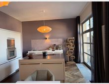 Modern kitchen interior with white built-in cabinets, a central island with a dark countertop, and a hanging pendant light against gray walls. Large floor-to-ceiling windows with dark curtains line one side, and a tall sculptural shelving unit stands near the window on a herringbone wood floor.